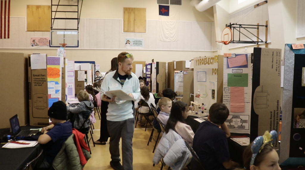 Man walks down aisle with project display boards on each side, and students sitting in front of each board. 