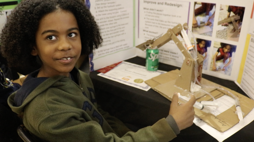 Student displays project board and cardboard mechanism