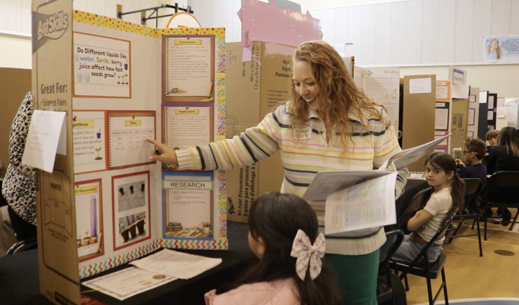 Woman points to project display board smiling and talking to girl seated in front of the board. 
