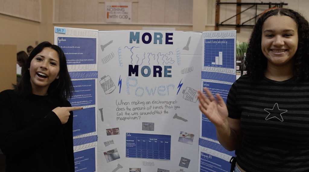 Two girls stand and pose in front of their project, " More Turns, More Power"