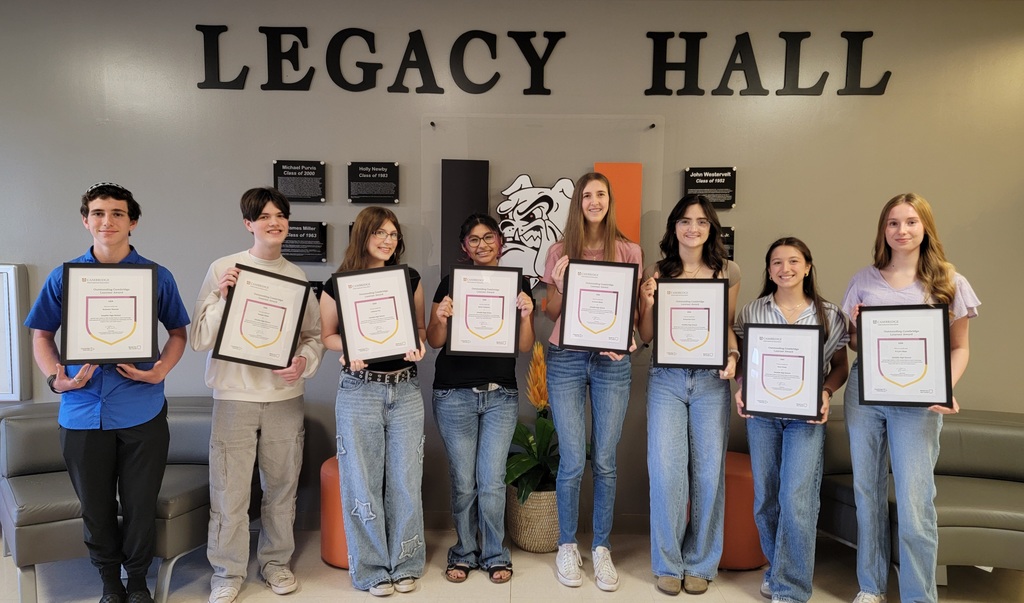 Nine students stand in a row inside Umatilla High School’s Legacy Hall, each holding a framed certificate recognizing academic achievement. Behind them on the wall are plaques and a large bulldog logo in the school’s orange and black colors. The students smile while posing with their awards, standing between two gray benches and decorative seating in the lobby area.