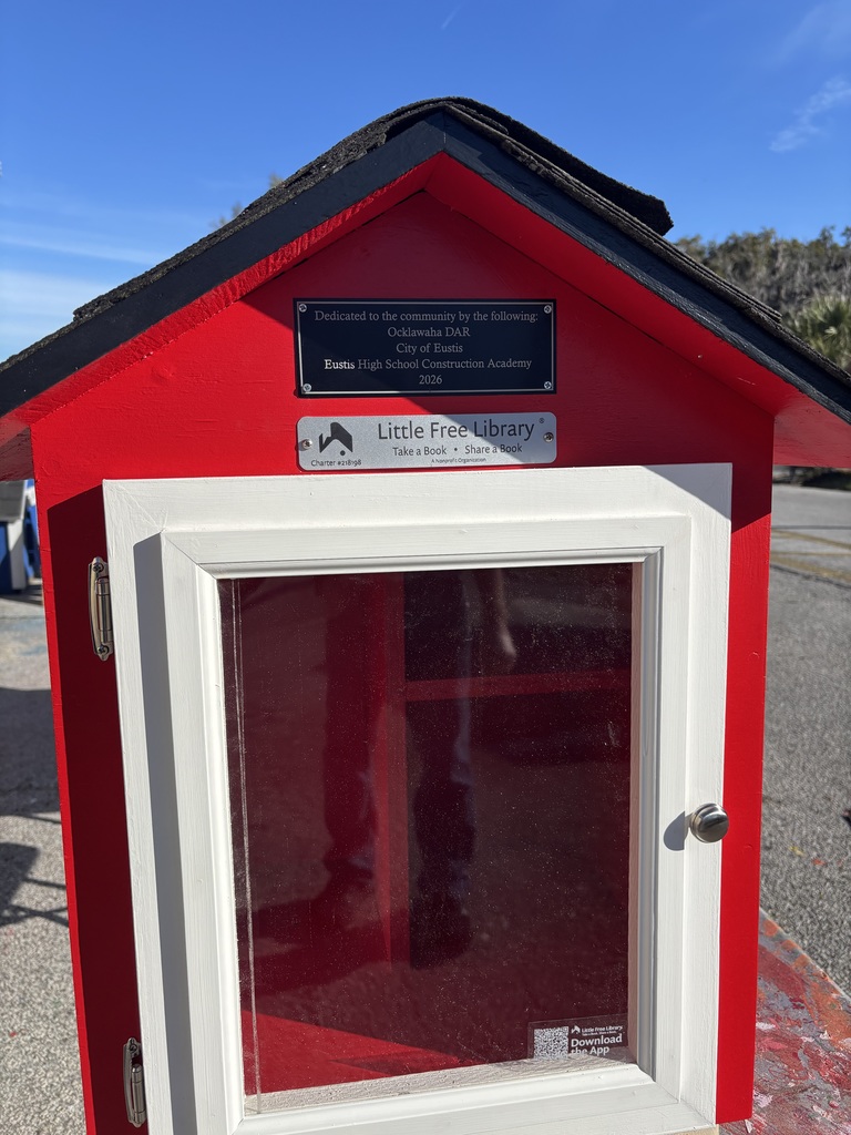 Closeup photo of a red tabletop house structure with a wide door. Signage says Little Free Library. Take a book, share a book. 