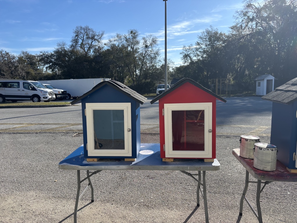 Photo of a blue and red tabletop house structure with a wide door. These will serve as little libraries. 