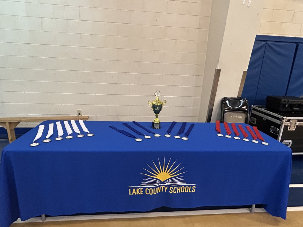 Red, white and blue ribbons and a trophy displayed on a tablecloth emblazoned with the Lake County Schools logo
