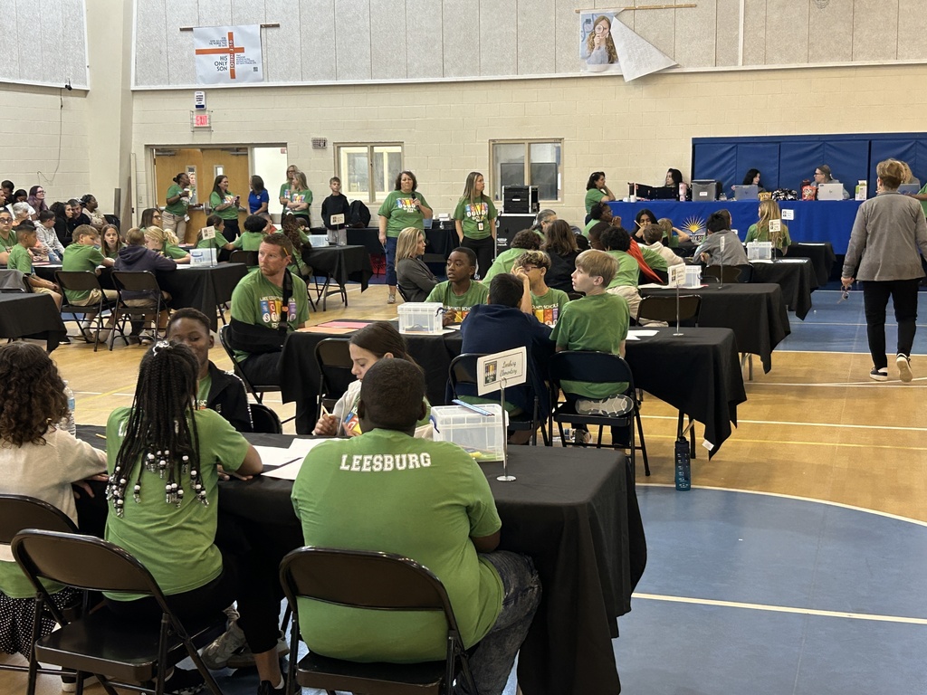 Students on gymnasium floor all wearing green t-shirts seated at tables with black tablecloths