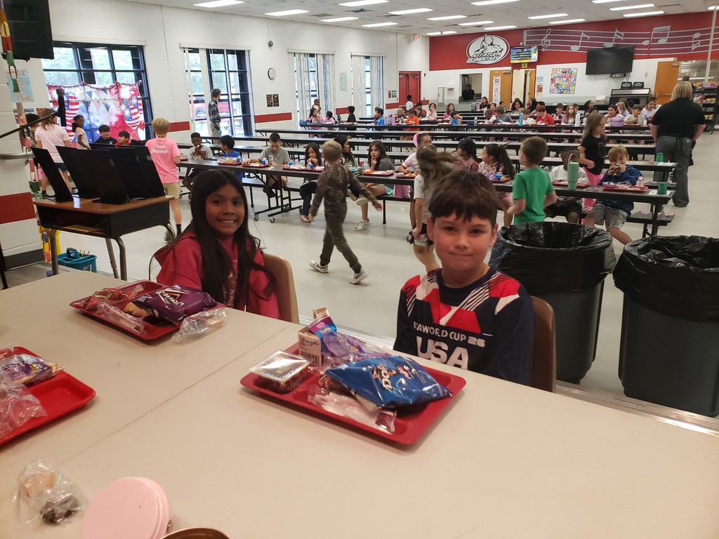 Busy school cafeteria at lunchtime with many children seated at long tables. In the foreground, a girl with long dark hair wearing a pink top smiles at the camera on the left; beside her a boy in a blue-red-white patterned shirt labeled "FAWORLD CUP 26 USA" looks toward the camera with a slight smile. Both have red lunch trays with snacks (a visible bag of Doritos on the girl's tray). Other students and an adult are visible in the background; large windows line the left side and a red-decorated wall with musical notes and a partial "AMERICAN…" banner is on the back wall.