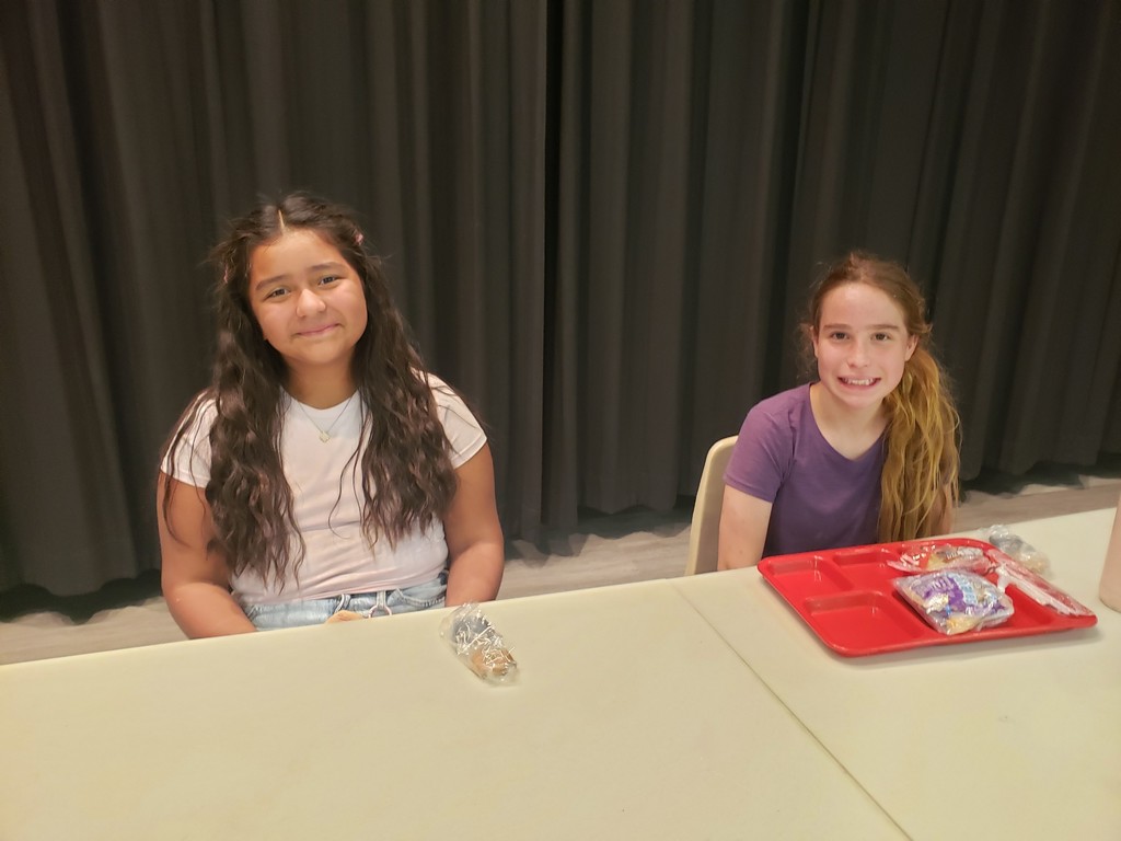Two girls sit at a white table smiling at the camera with a dark gray curtain behind them. The girl on the left has long dark wavy hair, wears a white T‑shirt and light jeans, and has a small packaged item on the table in front of her. The girl on the right has long lighter hair in a side ponytail, wears a purple T‑shirt, and has a red plastic tray containing several packaged items on the table before her.