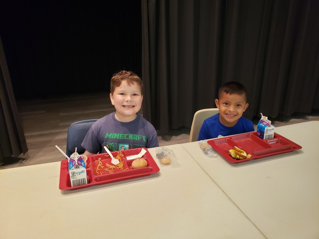 Two young boys sit side-by-side at a light-colored table, smiling at the camera against a dark curtain background. The boy on the left has light brown hair and wears a dark blue "MINECRAFT" T‑shirt; his red compartment tray holds noodles in sauce, a bread roll, a white plastic spoon, and a small round cookie or cracker on the table; a white-and-blue milk carton with a straw and a small plastic cup with a spoon sit beside the tray. The boy on the right has dark hair and wears a plain blue T‑shirt; his red tray contains several apple slices, and a white-and-blue milk carton with a straw and a clear plastic bag with two round cookies or crackers sit nearby.