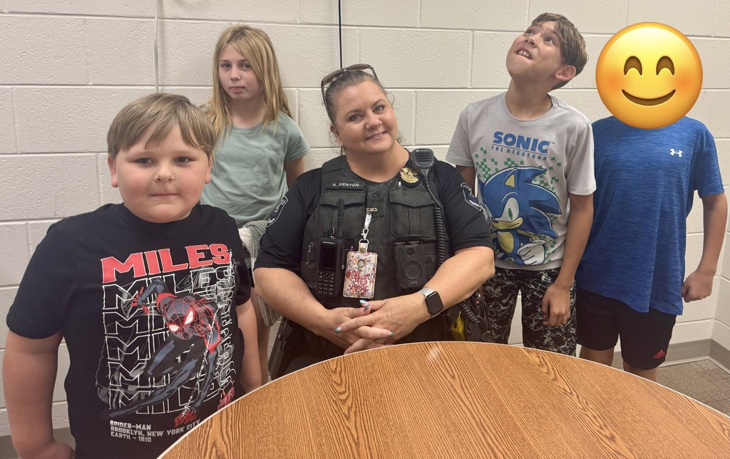 The students are making silly face expressions. Alt text: Smiling female police officer wearing a tactical vest labeled "A. DENTON," a Motorola radio, an Axon body camera, and a lanyard reading "QUEEN PONY," poses with four children in front of a pale brick wall. From left to right: a boy in a black shirt printed with "MILES, SPIDER-MAN, BROOKLYN, NEW YORK CITY, EARTH - 1610," a girl with long blonde hair in a light green shirt, a boy in a white "SONIC THE HEDGEHOG" shirt, and a boy in a blue Under Armour shirt (face later obscured by an emoji in another image). A wooden table is visible in the foreground.