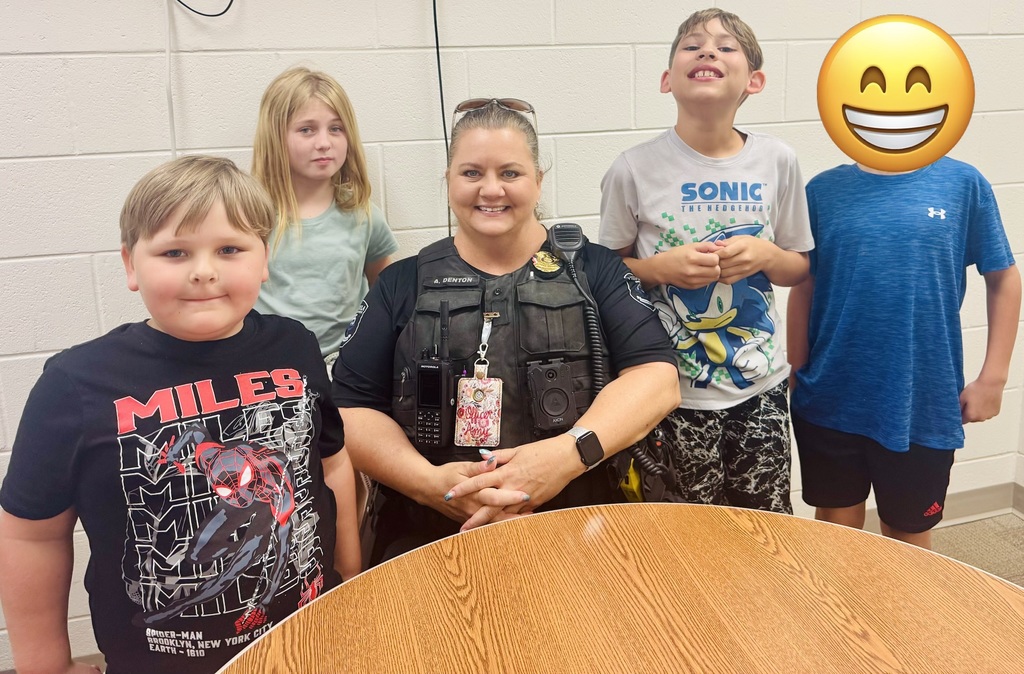 Alt text: Smiling female police officer wearing a tactical vest labeled "A. DENTON," a Motorola radio, an Axon body camera, and a lanyard reading "QUEEN PONY," poses with four children in front of a pale brick wall. From left to right: a boy in a black shirt printed with "MILES, SPIDER-MAN, BROOKLYN, NEW YORK CITY, EARTH - 1610," a girl with long blonde hair in a light green shirt, a boy in a white "SONIC THE HEDGEHOG" shirt, and a boy in a blue Under Armour shirt (face later obscured by an emoji in another image). A wooden table is visible in the foreground.