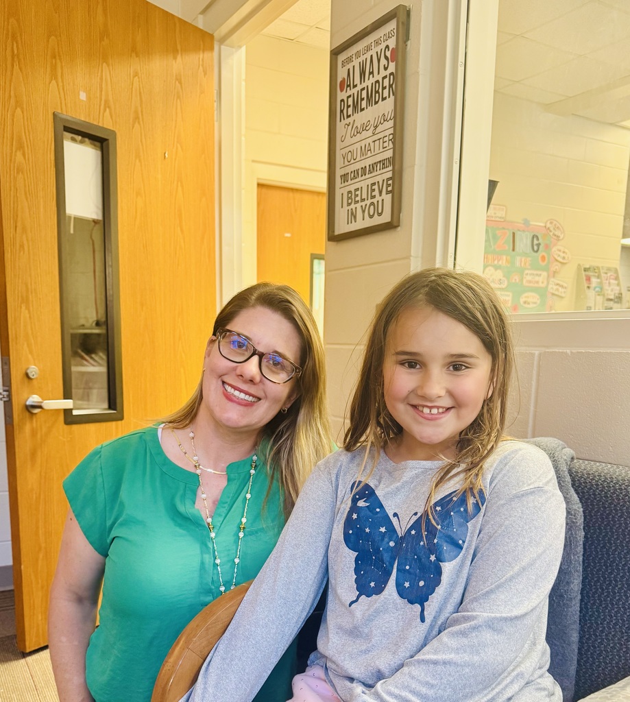 Medium close-up of an adult woman and a young girl seated side-by-side, both smiling at the camera. The woman (left) wears glasses, a green short-sleeved top, and a layered beaded necklace. The girl (right) wears a grey long-sleeved shirt with a large blue butterfly graphic decorated with white stars. Background shows an indoor school-like setting with a wooden door at left and a white wall with a framed motivational sign and a window into another room at right. The visible sign reads: "BEFORE YOU LEAVE THIS CLASS ALWAYS REMEMBER I love you YOU MATTER YOU CAN DO ANYTHING I BELIEVE IN YOU."