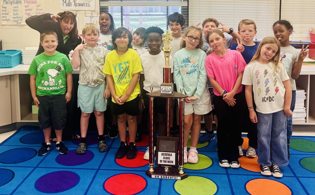 Group photo of an elementary classroom with teacher and students making silly faces. They are posing around a multi-tiered gold-and-red trophy labeled “ASTATULA CLASS OF THE WEEK” (also shows “GO BOBCATS!” on the trophy base). Image shows one adult female (likely the teacher) and a total of 24 elementary-aged children across the two photos (first photo: 13 children — 8 boys and 5 girls; second photo: 11 children). Most children are smiling and dressed casually in t-shirts and shorts or jeans. Background classroom details: blinds on windows, a whiteboard or poster titled “Math Resources,” and lists of multiplication and division vocabulary words (examples include multiplication: “each, twice, factor, multiplied by, equal groups”; division: “half, split, quotient, divisor, distribute, cut up, divided by, separate, shared equally, less than - fewer, take away - remain, left over - how many, difference - not”). Visible student shirt text includes “HERE FOR THE SHENANIGANS” (with a Snoopy graphic), “YETI,” “salt life / PARADISE,” and an “AC/DC” shirt with lightning-bolt graphics. One photo shows a blue rug with red, purple, and green circular patterns. Overall impression: a proud clas
