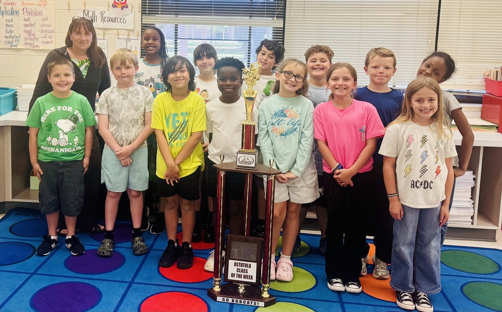Group photo of an elementary classroom with teacher and students posing around a multi-tiered gold-and-red trophy labeled “ASTATULA CLASS OF THE WEEK” (also shows “GO BOBCATS!” on the trophy base). Image shows one adult female (likely the teacher) and a total of 24 elementary-aged children across the two photos (first photo: 13 children — 8 boys and 5 girls; second photo: 11 children). Most children are smiling and dressed casually in t-shirts and shorts or jeans. Background classroom details: blinds on windows, a whiteboard or poster titled “Math Resources,” and lists of multiplication and division vocabulary words (examples include multiplication: “each, twice, factor, multiplied by, equal groups”; division: “half, split, quotient, divisor, distribute, cut up, divided by, separate, shared equally, less than - fewer, take away - remain, left over - how many, difference - not”). Visible student shirt text includes “HERE FOR THE SHENANIGANS” (with a Snoopy graphic), “YETI,” “salt life / PARADISE,” and an “AC/DC” shirt with lightning-bolt graphics. One photo shows a blue rug with red, purple, and green circular patterns. Overall impression: a proud clas