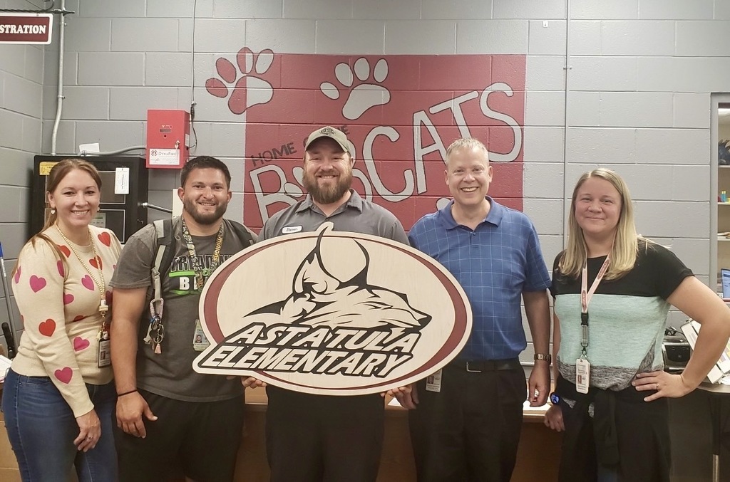 A group of five smiling adults (three men, two women) stand indoors in a row holding a large oval wooden sign reading “ASTATULA ELEMENTARY” with a stylized bobcat head logo. Behind them is a light gray cinder block wall with a red painted section showing a white mural of paw prints and the partial phrase “HOME OF THE BOBCATS.” Wall fixtures include a red fire alarm box labeled “DynaFIRE” and a partially visible “ADMINISTRATION” sign; one person wears a nametag reading “Steven.”