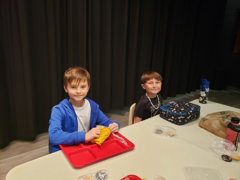 Two young boys sit at a cafeteria table in front of a dark curtain, both smiling at the camera. The boy on the left wears a blue hoodie over a white shirt and holds a yellow snack bag above a red tray. The boy on the right wears a black t-shirt and a white-and-blue beaded necklace; a patterned blue lunch bag, blue water bottle, brown paper bag, and a pink sports drink sit nearby. Several clear plastic bags with cookies are scattered on the table.