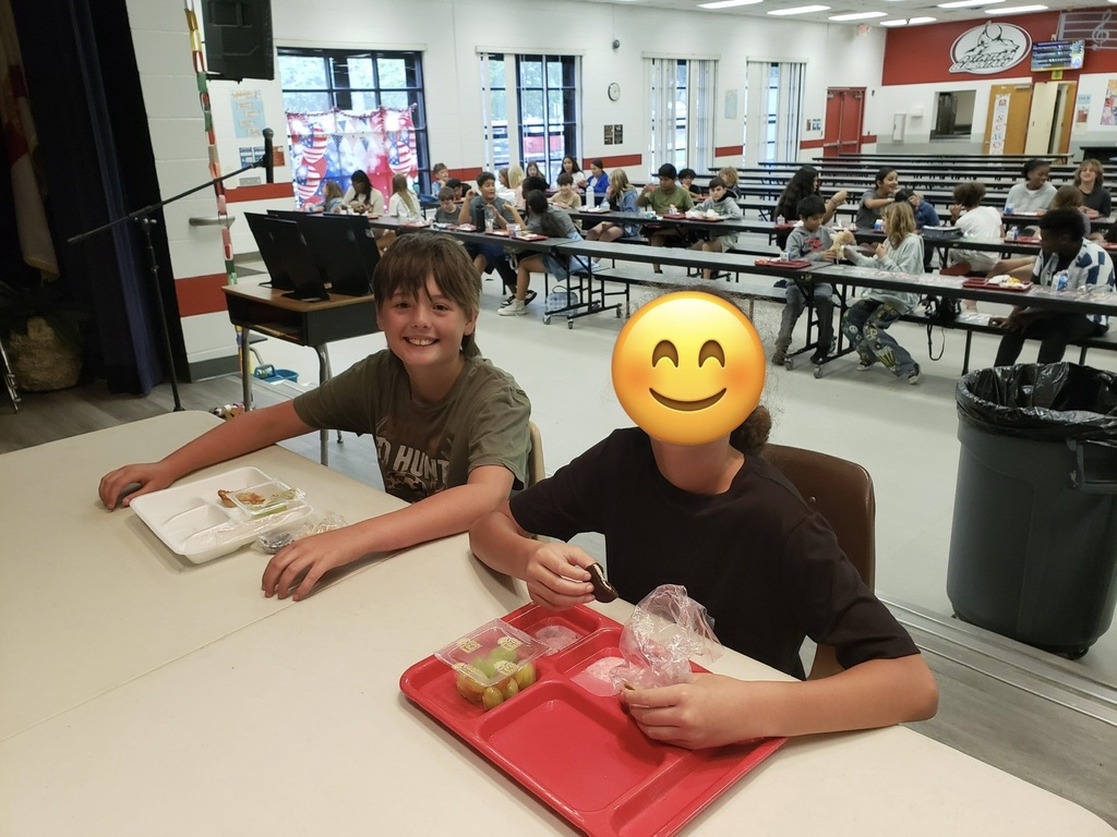 Image 2 alt text: A bustling school cafeteria filled with students seated at long tables. In the foreground two children sit at a table: a smiling boy on the left wearing an olive green shirt with “HUNT” partially visible, and a child on the right (face covered by a yellow smiley emoji) eating from a red tray that holds grapes and other items. Rows of students, a clock on the wall, large windows with red, white, and blue decorations, a stage area with flags and speakers, and a red wall logo/emblem are visible in the background.