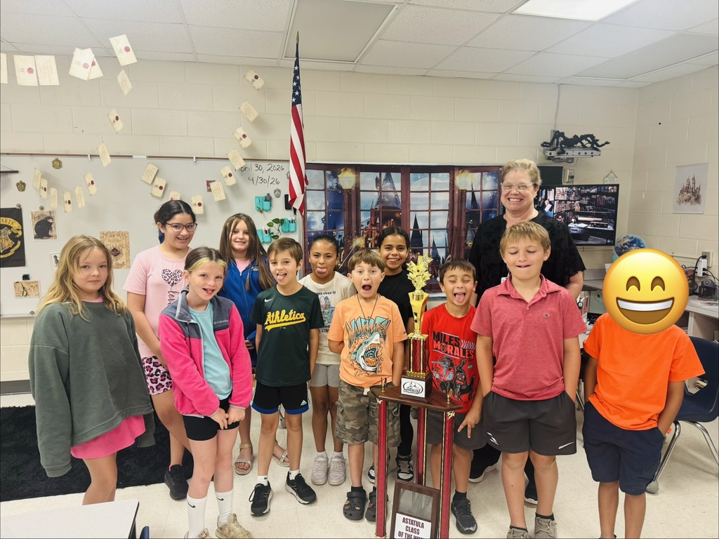 A smiling adult woman stands with ten elementary-age children in a classroom decorated with a Harry Potter theme — paper “flying” letters on a whiteboard, a Hogwarts-style crest, and a castle backdrop. An American flag is on the wall. A large gold trophy on a wooden stand sits in the center; the plaque shows partial text reading “ASTATULA CLASS OF THE WE.” Children wear colorful shirts (one green shirt reads “Athletica,” one red reads “MILES MORALES”). Several children make playful faces; the child at far right has their face covered by a yellow smiling emoji. Date text on the board reads “april 30, 2026 / 4/30/26.”