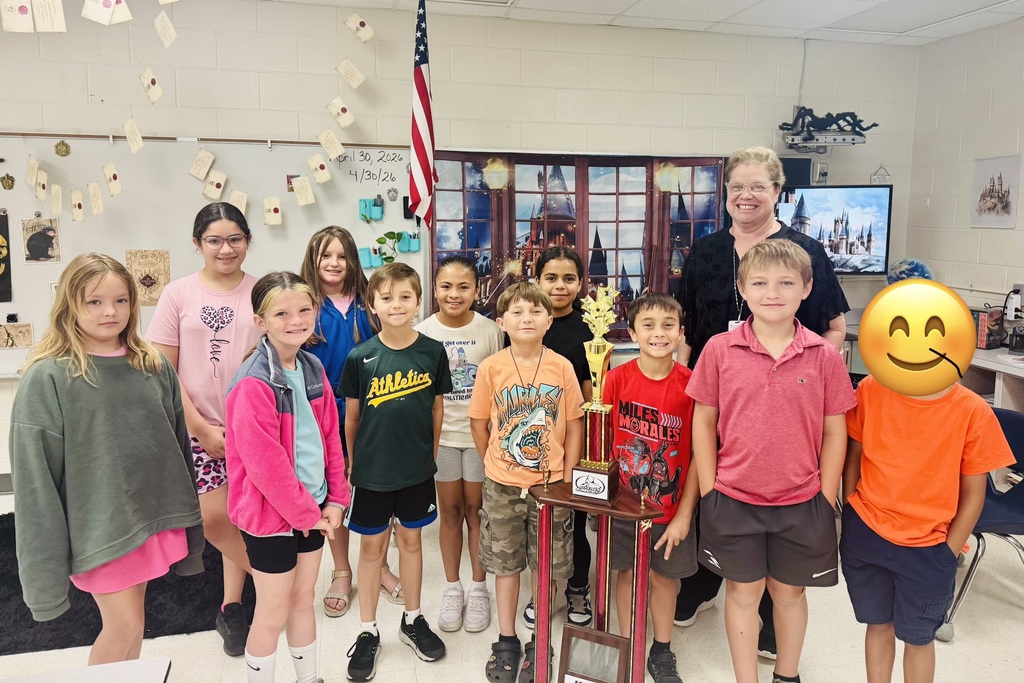 Ten elementary-age children and an adult woman pose in a Harry Potter–themed classroom with floating paper letters on a whiteboard and castle backdrops. A large trophy labeled “GENIUS” is held in front of the group. Visible shirt details include a green shirt reading “Athletics,” a pink shirt with the word “love” beneath a leopard-print heart, a white shirt reading “get over it / mad to mathema,” and a red shirt reading “MILES MORALES.” The adult stands behind the children, smiling. The child at the far right has their face obscured by a smiling emoji. Date on the board reads “April 30, 2026 / 4/30/26.”