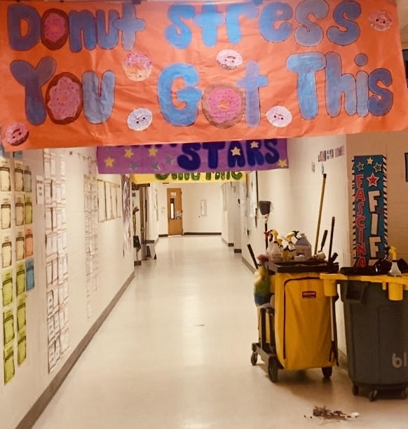 School hallway featuring a large orange banner reading “Donut Stress You Got This,” with donut images replacing some of the letter O’s. A purple banner below displays “STARS.” The left wall is covered with student papers or artwork. A yellow cleaning cart filled with supplies and a dark gray trash can with a yellow lid stand on the right; some small debris is on the floor nearby. A wooden door is visible at the far end of the hallway.