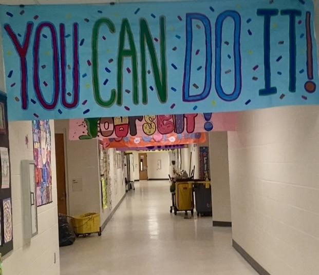 Long, well-lit school hallway with a large light-blue banner overhead reading “YOU CAN DO IT!” in hand-painted, colorful outlined letters (YOU purple, CAN green, DO IT! blue). A pink banner below shows the word “YOURSELF!” and additional banners hang further down. White walls are lined with student artwork on the left. A yellow mop bucket with wringer sits near a black trash bag in the foreground; a yellow cleaning cart and gray bin appear farther down the hall. Several closed wooden doors line the corridor.