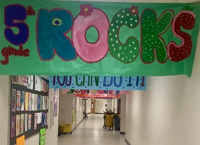 Decorated school hallway with a large green banner reading “5th grade ROCKS,” where letters in “ROCKS” are playfully styled (R blue, O pink with a flower, C and K green with white dots like cacti, S red with white dots like a strawberry). Below it hangs a blue banner that says “YOU CAN DO IT!” with colorful sprinkles. A bulletin board displaying many student works lines the left wall. The light-colored, reflective floor leads down the corridor past maintenance carts.