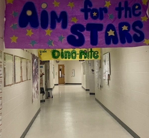 Bright school corridor with two hanging banners: a purple banner that reads “Aim for the STARS” decorated with yellow and teal stars, and a yellow-green banner below that says “grade Dino-mite.” Framed displays and a skeleton model are mounted on the left wall; a small sign reads “WELCOME ABST.” Papers and artwork are posted on the right wall. The polished floor reflects light and the hallway continues toward a wooden door in the distance.