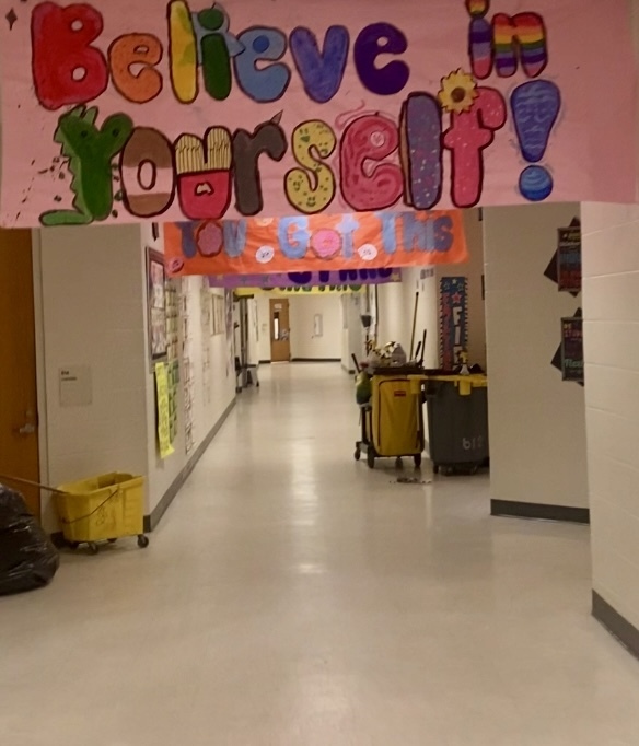 A bright school hallway decorated with two hand-painted banners. A large colorful banner across the ceiling reads “Believe in yourself!”; many letters are uniquely styled (the Y is a green dinosaur, the O is a donut, the U looks like french fries, the L includes a sunflower, and the exclamation mark is blue with white dots). Below it hangs an orange banner that says “You Got This.” Classroom door number 210 is visible on the left wall. Cleaning supplies (a yellow mop bucket and cart), a black trash bag, and a gray wheeled trash can are in the hallway; artwork and posters line the left wall. The corridor extends into the distance.