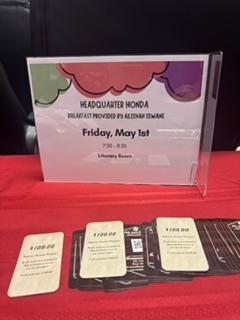 Tabletop display with a clear acrylic sign holder on a red tablecloth announcing “HEADQUARTER HONDA — BREAKFAST PROVIDED BY ALISHAH SEWANI” and event details (date, time, location). Promotional cards offering a $100.00 cash-back “Spouses House Program” for committing to buy a new vehicle are fanned in front of the sign. The setup conveys a sponsored breakfast and promotional offer.