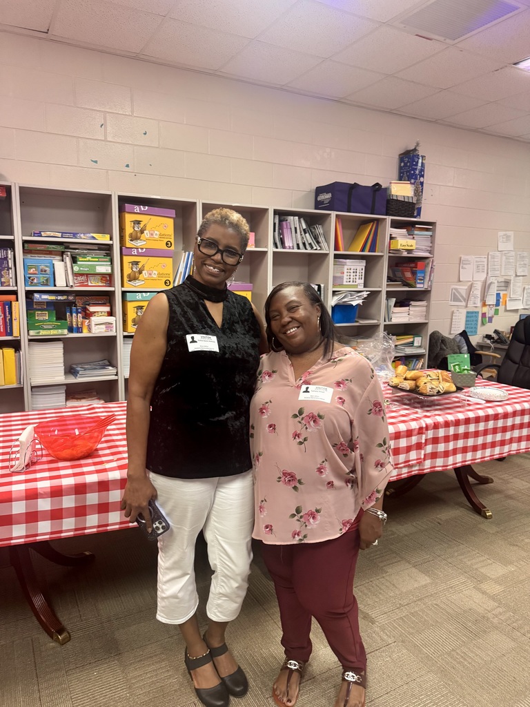 Two smiling women wearing "VISITOR" name tags stand side-by-side in a classroom or resource room with shelves of books, binders, and boxed materials behind them. The woman on the left has short light-colored hair, glasses, a black sleeveless top, and white capri pants; her name tag reads "VISITOR" and appears to say "Shelia." She holds a smartphone in her right hand. The woman on the right has dark hair, a pink floral blouse, and maroon pants; her name tag reads "VISITOR" and appears to say "Sylvia." In the foreground, a table with a red-and-white checkered tablecloth displays a clear red bowl of orange-colored items and a platter of pastries. Boxes on the shelves are labeled "Foundations" and "EasyScale." The scene suggests visitors delivering lunch for faculty and staff.