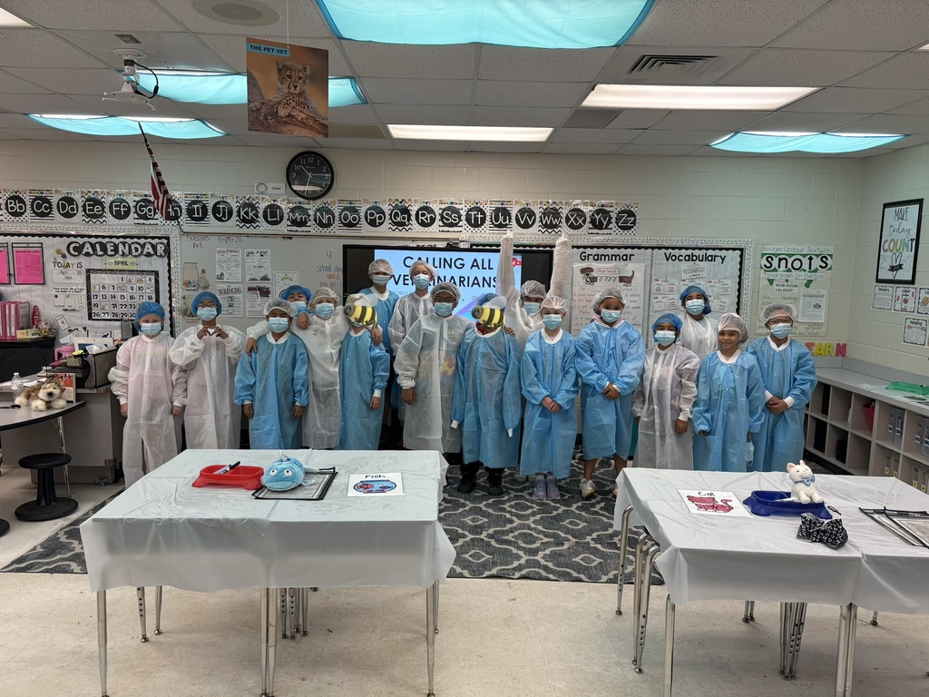 Group photo in a classroom showing about 16 children and a few adults dressed in light blue and white surgical gowns, masks, and hairnets, posing behind two tables with white tablecloths. Two children in the center wear large yellow-and-black striped bee masks. Classroom walls display educational posters including a large cheetah poster titled "THE PET VET," an alphabet line, a calendar showing "April," and a digital screen reading "CALLING ALL VETERINARIANS." Foreground tables hold pet-related props: a red tray with tools, a blue stuffed toy, drawings labeled "Fish" and "Cat," a blue pet bowl, and a white cat figurine. A stuffed husky toy sits on a nearby desk. The scene suggests a veterinary-themed school activity.