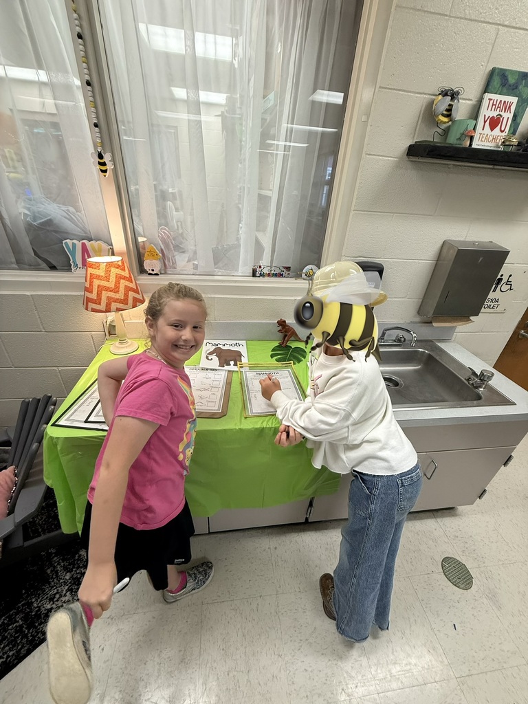Two children at a green-covered table in a classroom. A blonde girl in a pink shirt and black shorts smiles at the camera while holding a shoe; another child wearing a bee-themed head covering writes on a worksheet. The table displays papers titled "Mammoth" with illustrations and a small mammoth/dinosaur figurine. Behind them are a window with white curtains, a shelf with a "Thank You Teacher" sign and bee figurine, and a sink area with a sign indicating "510A TOILET" and an accessibility symbol. The scene suggests a themed classroom activity about mammoths.