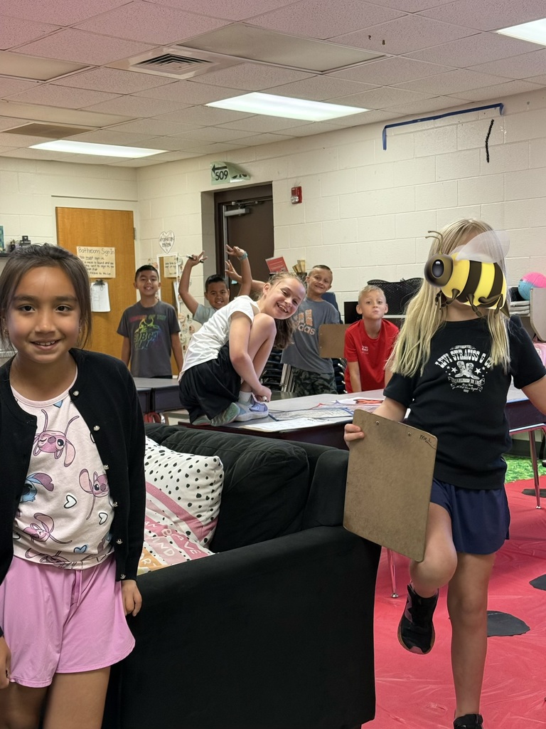 Group of elementary-aged children posing in a classroom. In the foreground, a dark-haired girl wearing a black cardigan and pink Lilo & Stitch t-shirt smiles at the camera; to her right a girl holding a clipboard is partially obscured and her face is covered by a bee emoji. Five other children pose in the background, with one squatting on a desk. The room has white brick walls, fluorescent lights, a wooden door, and a red floor covering with black circles. Visible wall text includes a bathroom sign "509" and brand names like "Levi Strauss & Co." and "adidas."