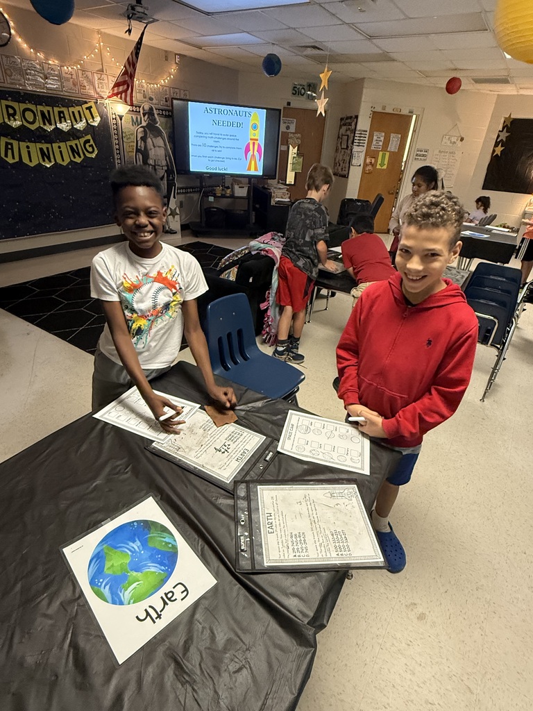 Bright, decorated classroom where children participate in a space-themed activity. Two boys stand at a table covered with a black cloth, smiling and holding markers while working on activity sheets showing Earth and space icons. The large screen behind them reads "ASTRONAUTS NEEDED!" with instructions about completing 10 challenges to earn the title of astronaut. Ceiling decorations include hanging stars and balloons; a banner on the back wall reads "ASTRONAUT TRAINING." Other students and space-themed posters (including a character resembling Darth Vader) are visible in the background.