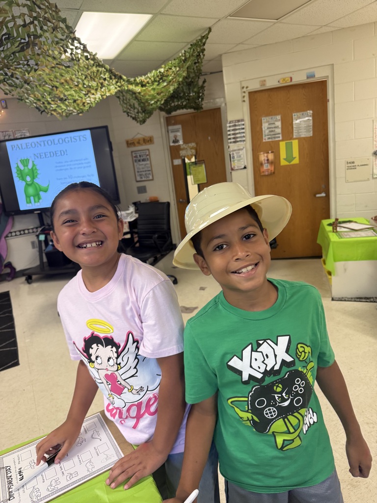 Two smiling children stand behind a green-covered table in a dinosaur-themed classroom. The girl on the left wears a light pink Betty Boop shirt; the boy on the right wears a green Xbox-controller shirt and a light-colored pith helmet. A worksheet titled "DINOSAUR DAY" is on the table. A large TV screen in the background displays "PALEONTOLOGISTS NEEDED!" with an image of a green triceratops-like dinosaur and a promotion for a summer "Dinosaur Day" with fossil-related activities. Classroom decorations include camouflage netting on the ceiling and various motivational posters on the walls.