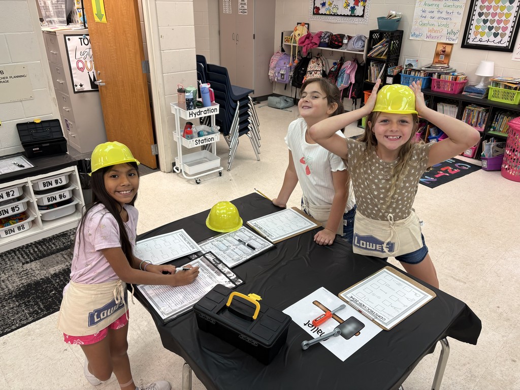 Three young girls in a classroom dressed for a construction-themed activity: each wears a yellow hard hat and a beige apron with a Lowe’s logo. They stand around a table draped in black, smiling and working with papers, clipboards, a small toy toolbox, and plastic tools (hammer and wrench). One girl writes on a sheet while another adjusts her hard hat. Classroom background shows labeled storage bins, educational posters, a white rolling cart labeled “Hydration Station,” stacked chairs, and visible signage with headings like “CONSTRUCTION ZONE,” “ASSEMBLE CHALLENGE,” and a poster about answering questions. The scene conveys a hands-on learning activity focused on building and problem-solving.