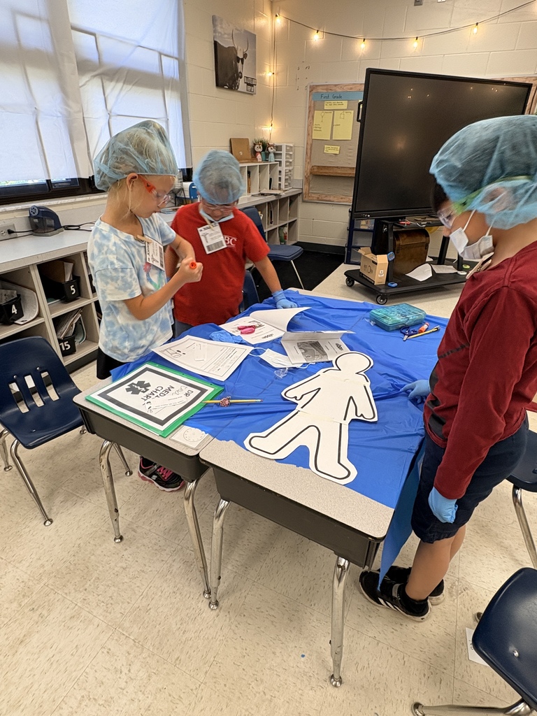 Three children wearing blue surgical caps and gloves sit at two desks pushed together under a blue "surgical" drape. A large white paper silhouette of a person is on the table with scissors, pencils, and worksheets nearby. A green-framed document labeled "DR MEDICAL CHART" rests on an adjacent desk. Classroom cubbies and a display screen are visible behind them. The scene shows students engaged in a themed text-feature activity using medical charts.