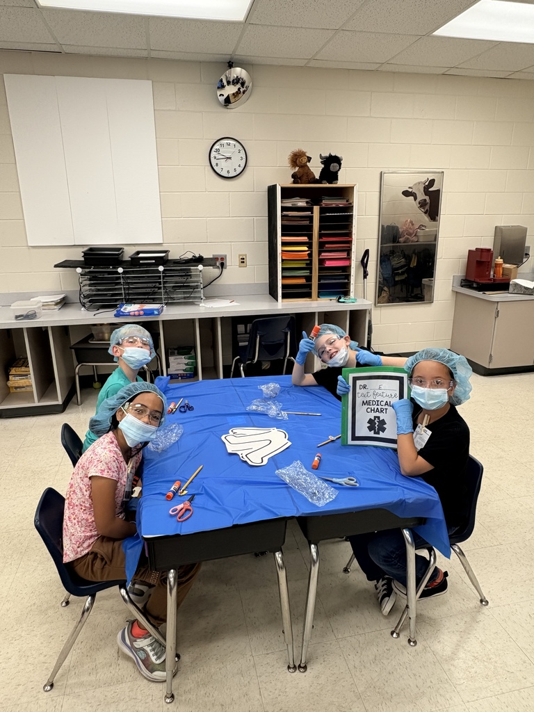  Four children at a table covered with a blue tablecloth wearing light-blue surgical caps, safety glasses, and gloves. Craft supplies, a white paper cutout resembling an internal organ, and clear plastic pieces are on the table. One child holds a green binder labeled "MEDICAL CHART." Classroom shelves, stuffed animals, a wall clock, and a whiteboard are visible in the background. The image suggests a hands-on "text feature surgery" activity using medical-chart props.