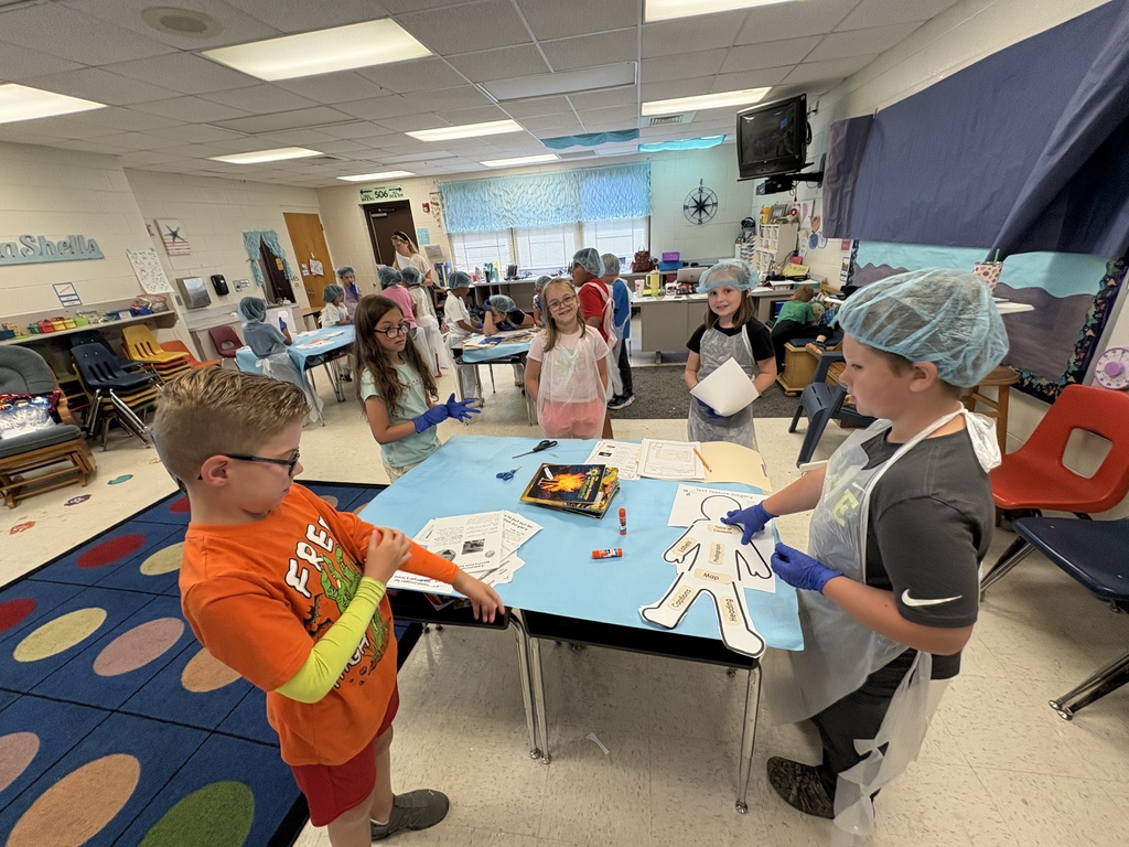 Bright classroom with elementary students wearing blue hairnets, plastic aprons, and gloves during a hands-on activity. Two boys stand at a table with a paper cutout of a human figure labeled with terms; other students work at similar stations in the background. Wall decor includes a "SeaShells" sign and a compass rose. A visible paper title reads "Text Feature Surgery," indicating the group is identifying and labeling nonfiction text features in a mock medical/surgical format.