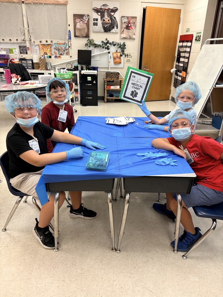 Four young children seated around two desks covered with a blue tablecloth, dressed as medical professionals in blue surgical caps, face masks, and some wearing goggles and gloves. One child holds a framed sign reading "MEDICAL CHART" while craft supplies and paper cutouts lie on the table. Classroom walls show animal artwork and a whiteboard. The scene depicts a role-play activity where students examine a "medical chart" as part of a text-feature lesson.
