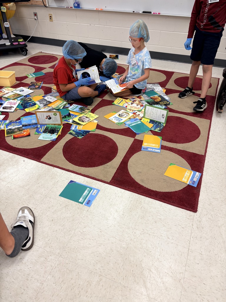 Several children and an adult on a red-and-beige patterned rug wearing blue hairnets and gloves, looking through a spread of books. Two children sit and hold open books while an adult lies on the rug wearing a hairnet. Colored sheets with labels and many science- and nature-themed book titles are scattered around. The activity appears to combine book exploration with the "Text Feature Surgery" theme.