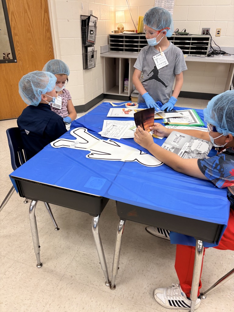 Four elementary students gathered around a table covered with a blue plastic tablecloth, wearing blue surgical hairnets, masks, and gloves; one child also wears red-framed safety glasses. A large white paper outline of a torso is on the table along with worksheets and a glue stick. A shelf sign reading "BEST TEACHER EVER" and a folder labeled "MEDICAL" are visible in the background. The image conveys a collaborative, medical-themed lesson about parts and text features.