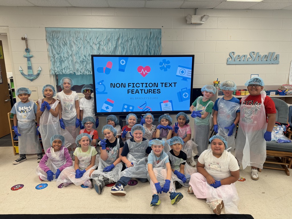 Group photo of about 20 students in Ms. Bradley’s classroom, all wearing light-blue bouffant caps, clear plastic aprons, and blue gloves. A large screen behind them displays "NON FICTION TEXT FEATURES — MS. BRADLEY'S CLASS" with medical-themed icons (microscope, first-aid kit, heart monitor). Classroom decorations include seaside-themed elements and a "SeaShells" sign. The scene shows a themed, schoolwide "Text Feature Surgery" activity where students explore nonfiction text features in a playful medical context.