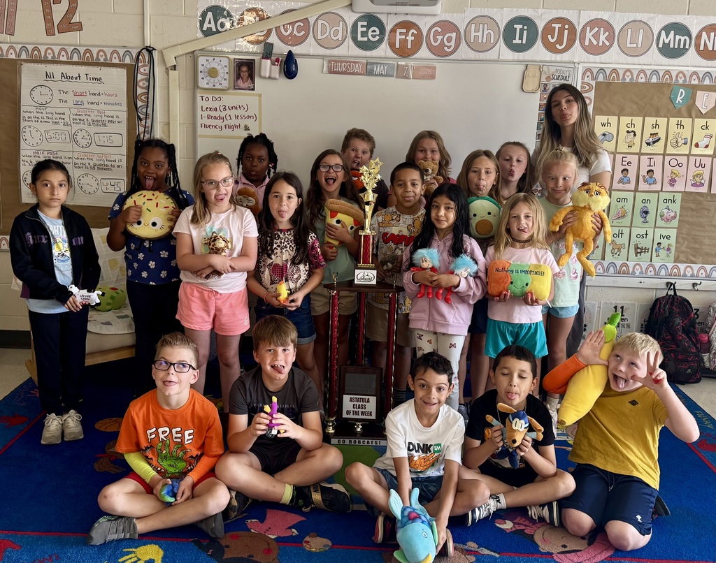 Bright classroom group photo of roughly 18 elementary students with one adult teacher. Students pose on a blue patterned carpet, many holding colorful plush toys; one child holds a green pillow reading "Nothing is IMPOSSIBLE!" A large gold-and-red trophy is centered in the group. The whiteboard behind them shows an alphabet line, the date "THURSDAY, MAY 7, 2026," and a "TO DO" list (Lexia, I-Ready, Fluency flight). Posters include "All About Time" with clock examples and phonics charts. Several students make playful faces; overall atmosphere is celebratory and cheerful.