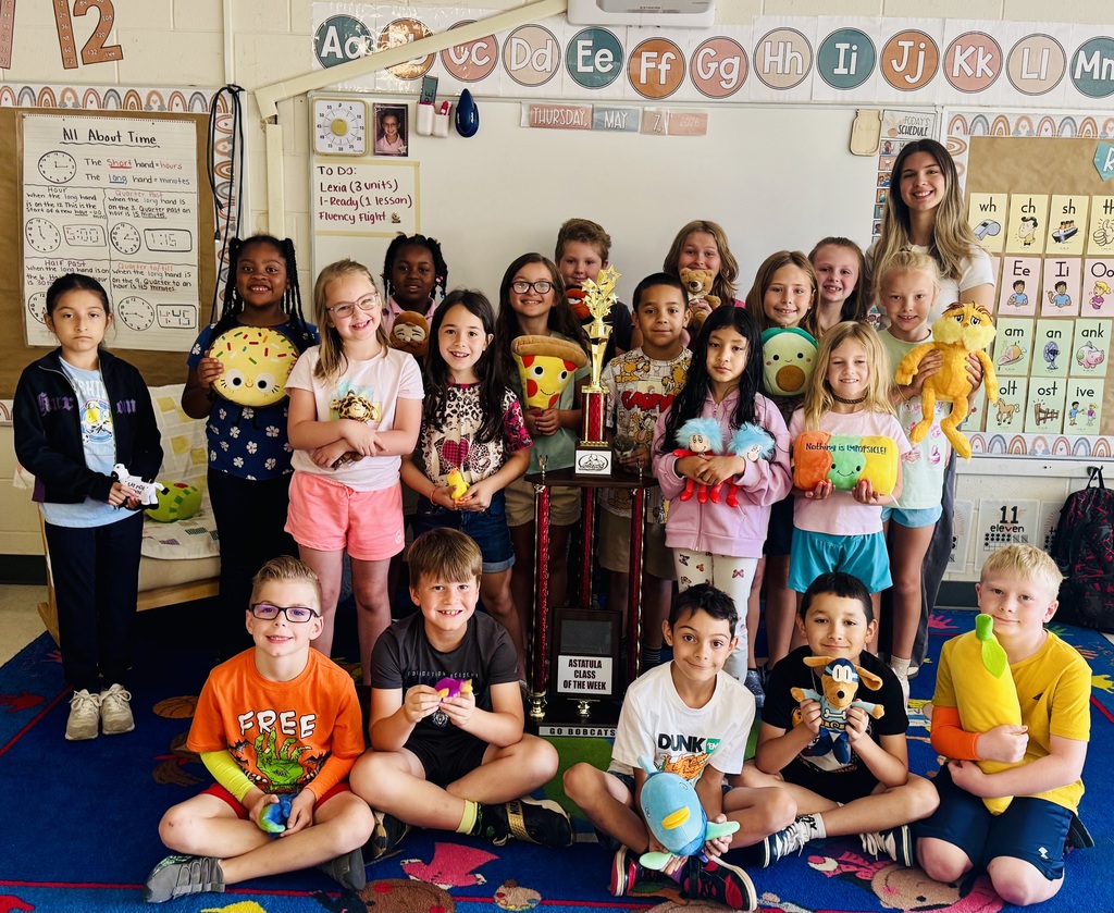 Class photo of about 19 elementary students and one adult teacher standing and sitting on a colorful classroom rug. A large trophy and a smaller trophy sit near the center. Many children hold plush toys (donut, pizza slice, avocado, Thing 1 & Thing 2, banana, various animals). Whiteboard behind them displays an alphabet line, the date "THURSDAY, MAY 7, 2026," a "TODAY'S SCHEDULE!/TO DO" list (Lexia, I-Ready, Fluency flight), an "All About Time" poster with clocks, and a phonics chart. A floor mat reads "ASTATULA CLASS OF THE WEEK GO BOBCATS." The scene celebrates Miss Exline’s class being Class of the Week.  Exline 2.jpeg — Alt text: Bright classroom group photo of roughly 18 elementary students with one adult teacher. Students pose on a blue patterned carpet, many holding colorful plush toys; one child holds a green pillow reading "Nothing is IMPOSSIBLE!" A large gold-and-red trophy is centered in the group. The whiteboard behind them shows an alphabet line, the date "THURSDAY, MAY 7, 2026," and a "