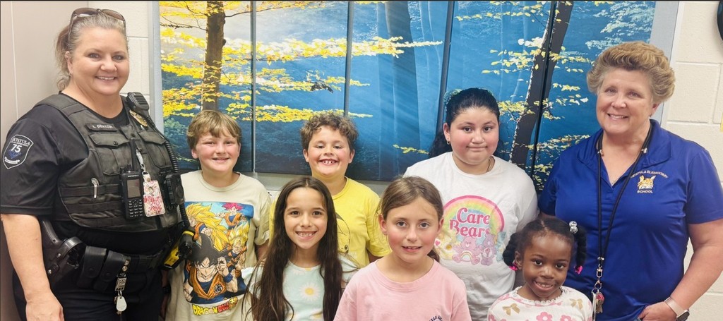 Small group photo of five smiling children and two adult women standing in a school hallway with framed tree artwork on the wall behind them. The woman on the left is a police officer with a vest labeled "L. DENTON"; the woman on the right wears a blue polo with an Astatula Elementary School bobcat logo. Children wear casual shirts, including "Dragon Ball Z" and "Care Bears" graphics. The image shows students honored at a special Lunch Bunch with school staff and a school resource officer.
