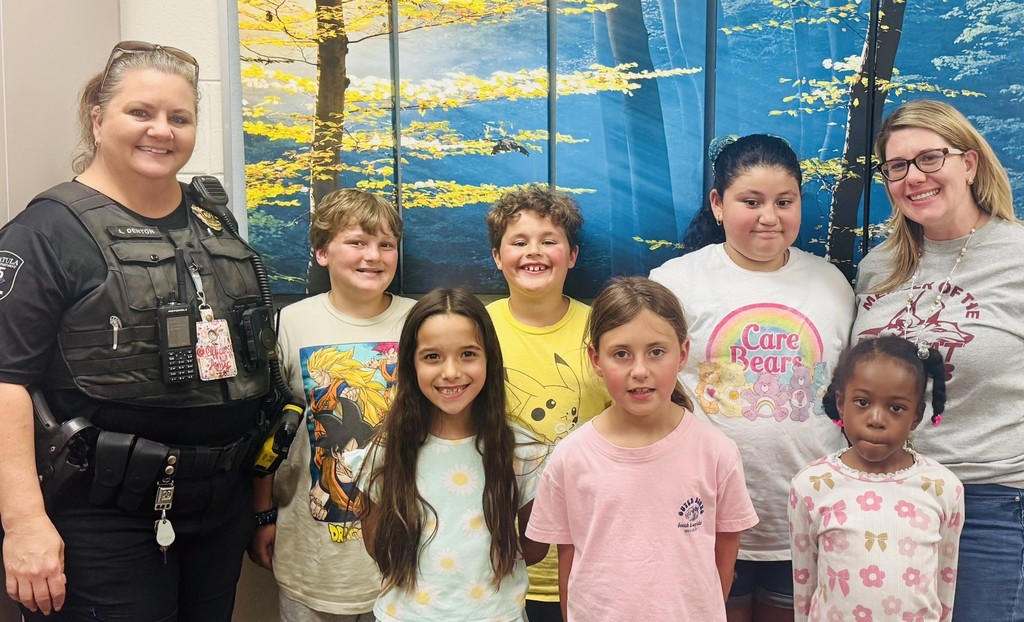 Group photo of seven smiling children with two adult women standing indoors in front of murals showing yellow-leaved trees and a blue sky. On the left, a female police officer wears a black uniform vest with a name tag reading "L. DENTON" and a visible police patch; she has sunglasses pushed up on her head. On the right, an adult in glasses wears a light gray t-shirt and beaded necklace. Children wear casual graphic t-shirts (including Dragon Ball, Pikachu, Care Bears, and Outer Banks) and pose in two rows—three in front and four behind. The scene captures a celebratory Lunch Bunch moment recognizing students for positive behavior.