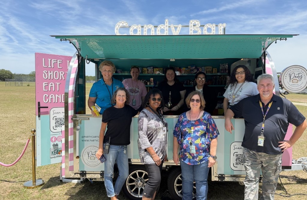 Group photo of eight smiling adults standing in front of a pink-and-teal "Candy Bar" truck parked on a grassy field. The truck features prominent signage reading "Candy Bar," branding for Party Carts & Co., QR codes, and social handles @PARTYCARTCO and  www.partycarts.co(opens in new tab) . A gold stanchion and pink rope mark the truck entrance; the sky is blue with scattered clouds.