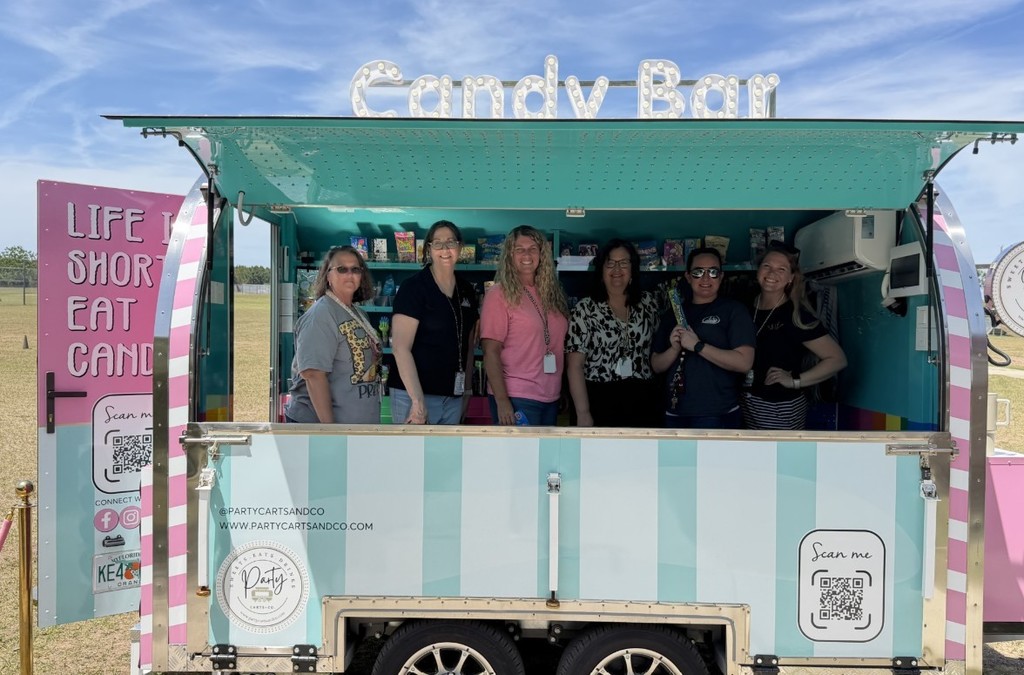 Six women stand inside an open, striped pink-and-teal candy truck smiling and posing while holding candy. The truck interior shows shelves stocked with candy and an illuminated "Candy Bar" sign on the roof. Exterior panels display messages like "LIFE IS SHORT EAT CANDY," social media icons, QR codes, and @PARTYCARTSANDCO with the website. The truck is parked in a grassy field on a sunny day.