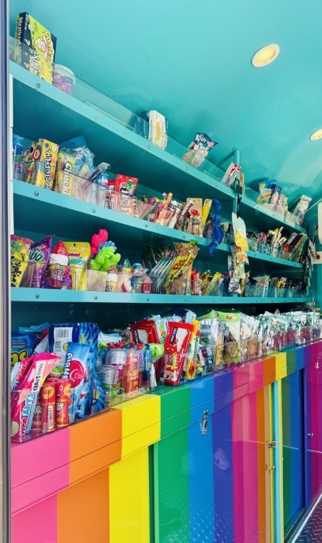 Interior view of a colorful candy cart showing multiple turquoise shelves filled with packaged sweets and snacks. Visible items include Airheads, Skittles, Warheads, Chupa Chups, Push Pop, and small novelty toys. The counter below is piled with assorted candy bags; the cabinet fronts feature rainbow vertical stripes. Two round recessed lights are visible in the turquoise ceiling.