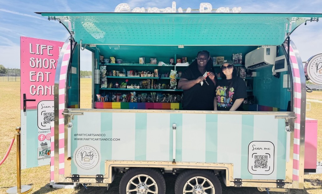 Woman inside a colorful mobile candy cart stocked with shelves of packaged sweets and snacks. The cart interior is aqua-teal with a rainbow-striped lower trim. Exterior signage shows "LIFE IS SHORT EAT CANDY," a large QR code labeled "Scan me," and @PARTYCARTSANDCO with the website. The woman wears a black "CANDY CREW" shirt and sunglasses; the cart is parked outdoors on a sunny day.